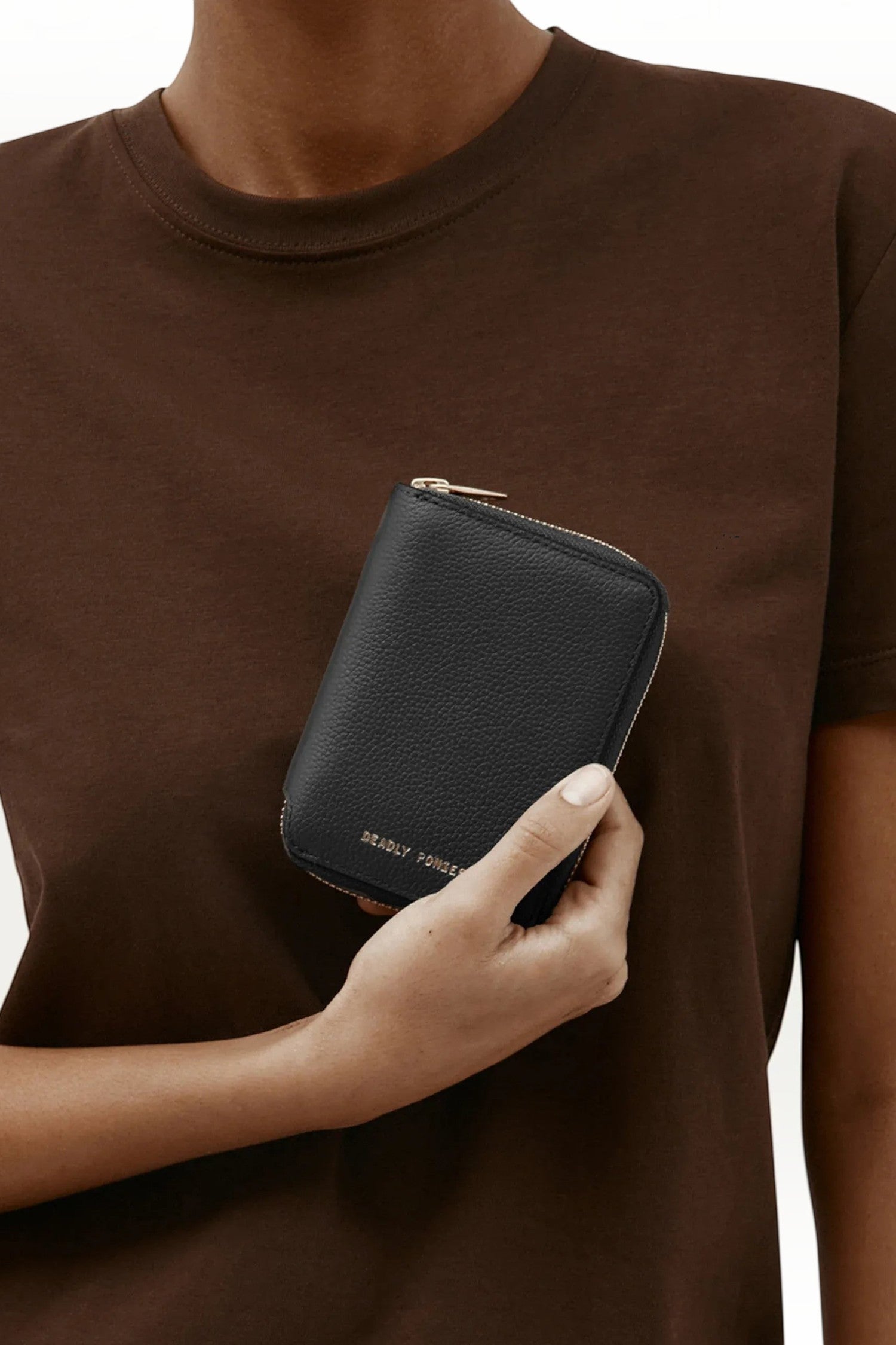 ELSHAM BOUTIQUE black grained calf leather mini wallet with brass zip closure held in a hand against a brown shirt.