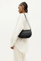 ELSHAM BOUTIQUE black calf nappa mini sling bag with brass hardware worn over the shoulder by a model in a white shirt