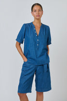Model wearing an ELSHAM BOUTIQUE short-sleeve chambray linen blouse with a V-neck, button placket, and puffed sleeves.
