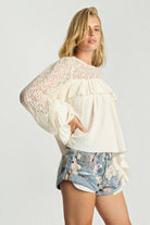 Model wearing ELSHAM BOUTIQUE ivory cotton blouse with lace sleeves, frill detailing, and a high neckline.
