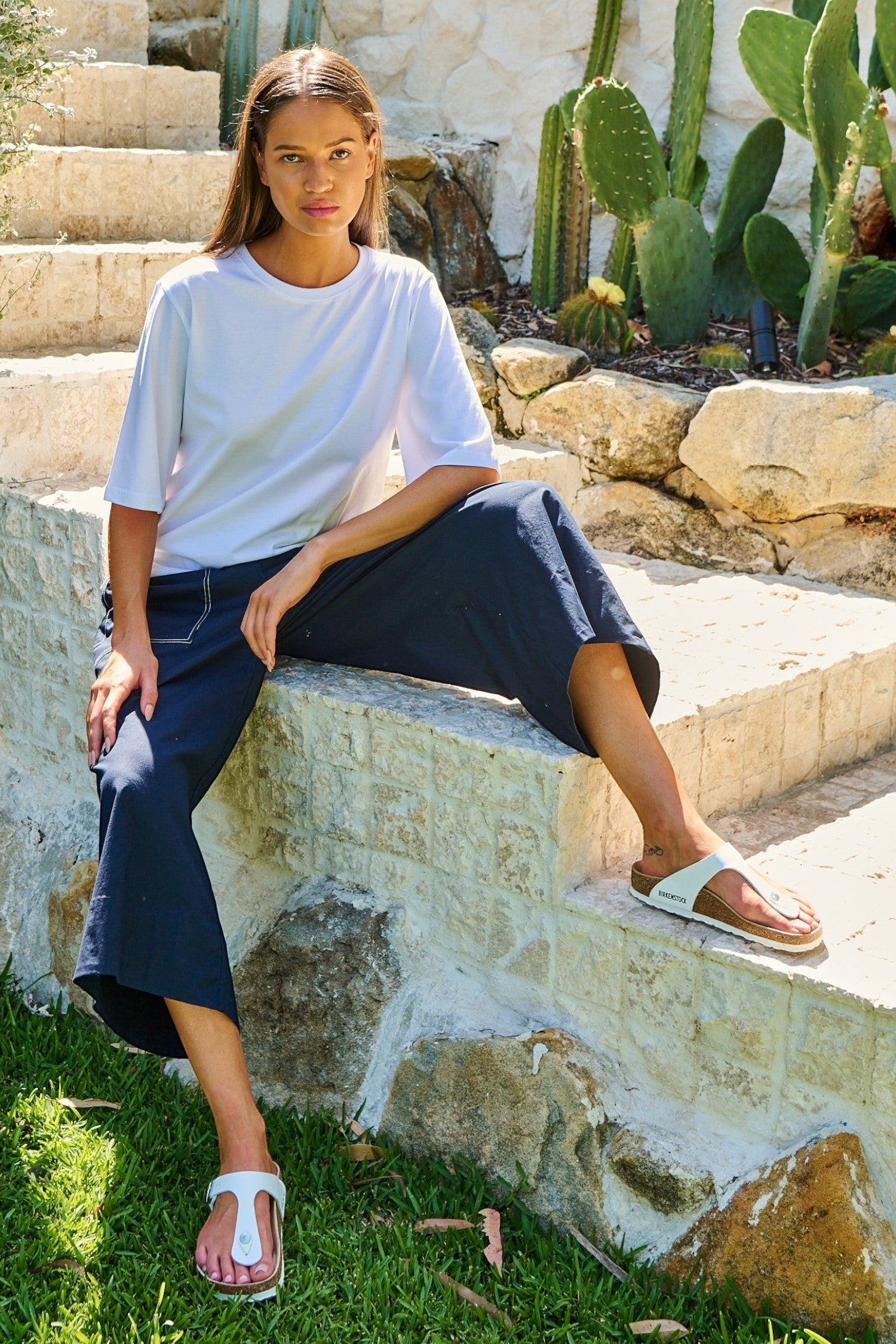 ELSHAM BOUTIQUE white cotton drop shoulder crew neck top worn by a model sitting on stone steps in a garden