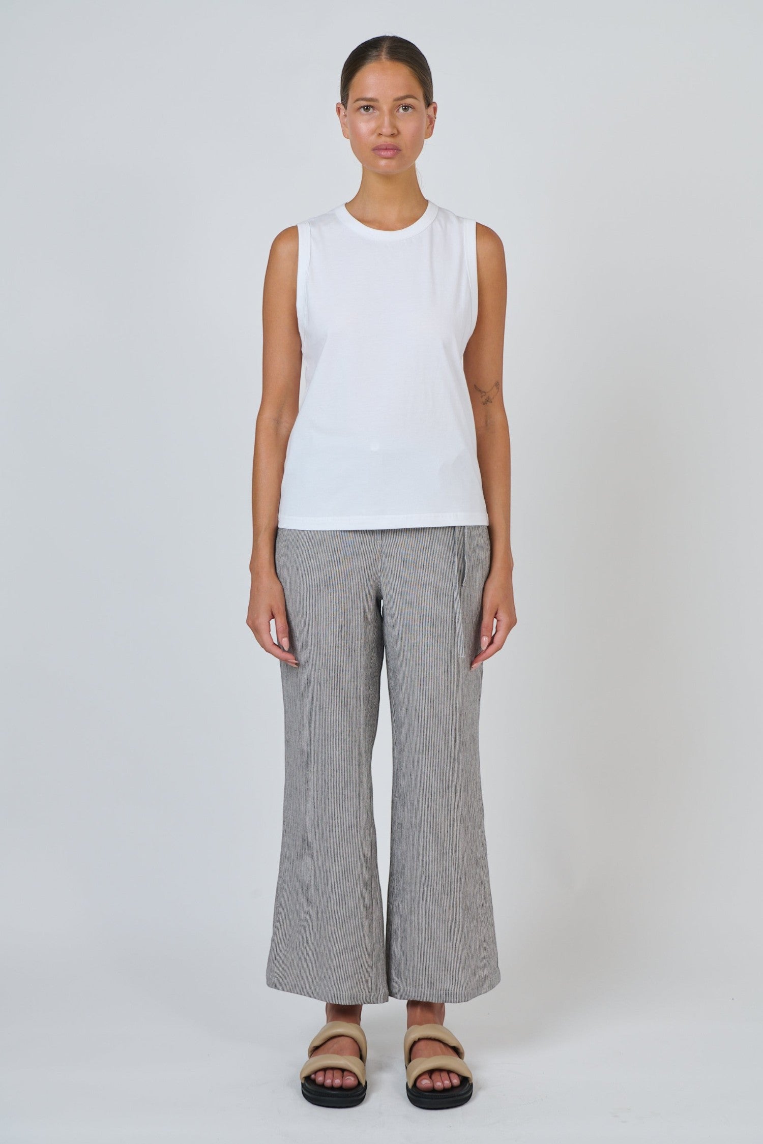 ELSHAM BOUTIQUE white cotton sleeveless top with a round neckline worn by a model with grey trousers and sandals.